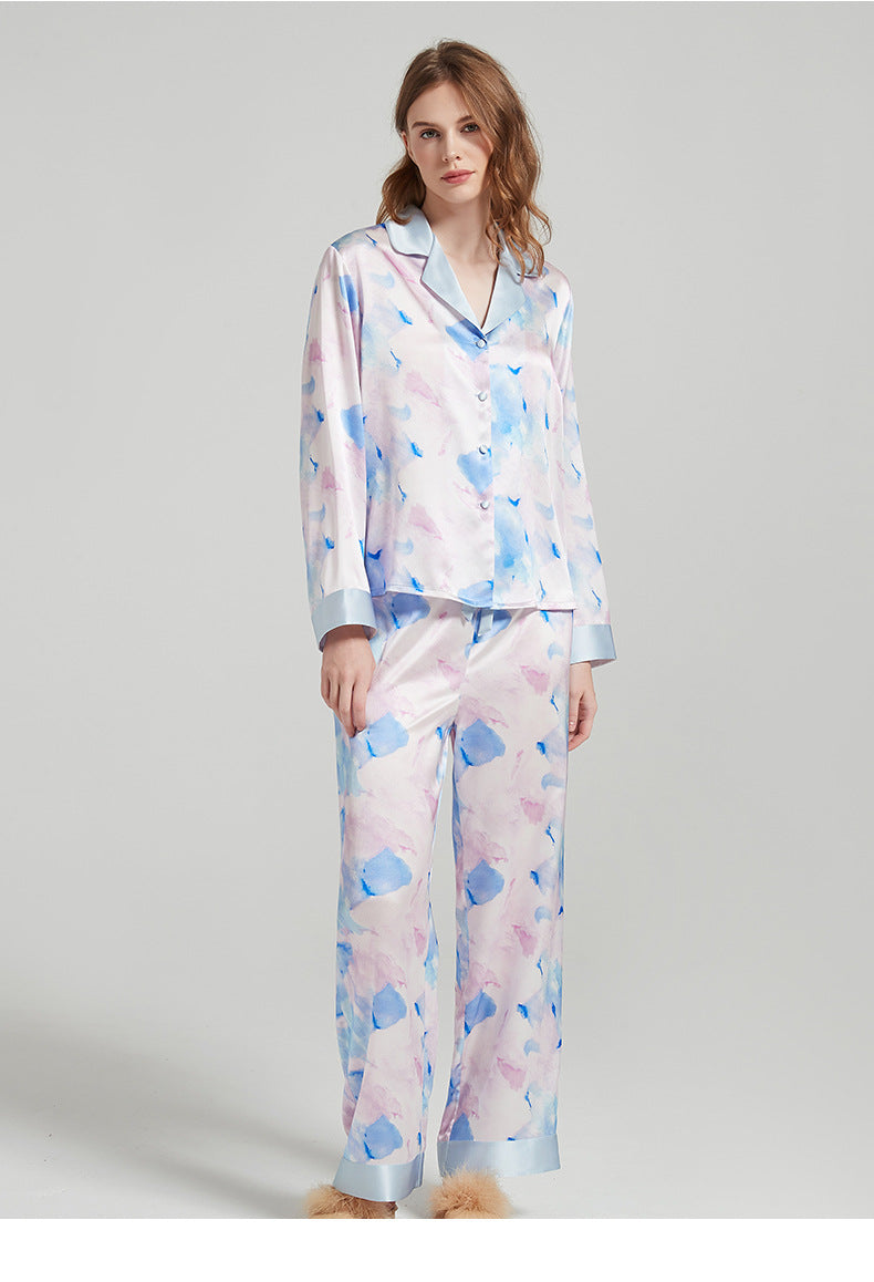 A model in a watercolor-printed lavender and blue silk pajama set with light blue piping, paired with tan fluffy slippers.