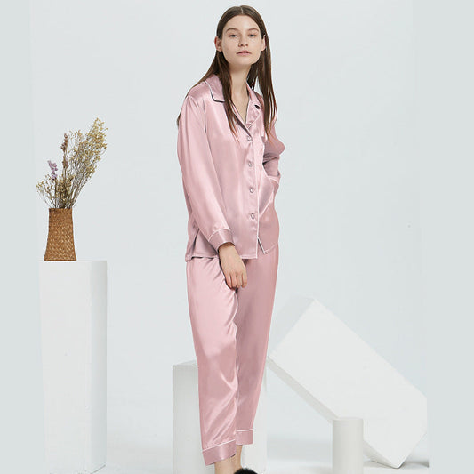 Woman standing in a studio wearing a pink satin pajama set with a button-front top and matching pants, beside white geometric blocks and a woven vase with dried flowers