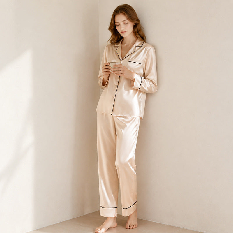 A woman standing barefoot in a corner wearing a light apricot silk pajama set with black piping, holding a small cup against a neutral wall