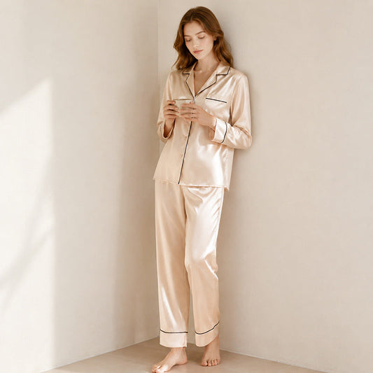 A woman standing barefoot in a corner wearing a light apricot silk pajama set with black piping, holding a small cup against a neutral wall