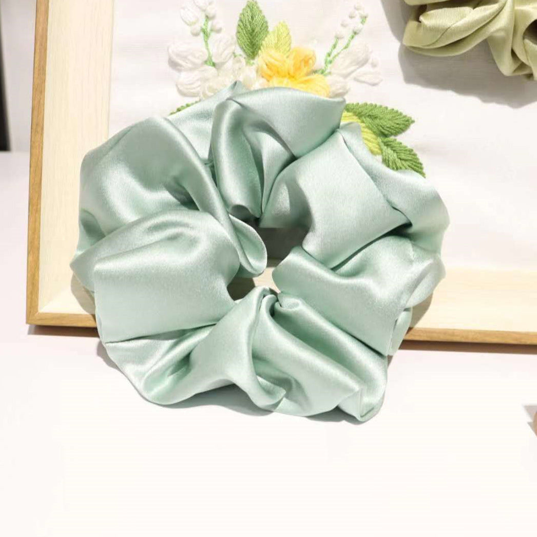 Mint green mulberry silk scrunchie with ruched folds resting against a wooden-framed floral embroidery piece