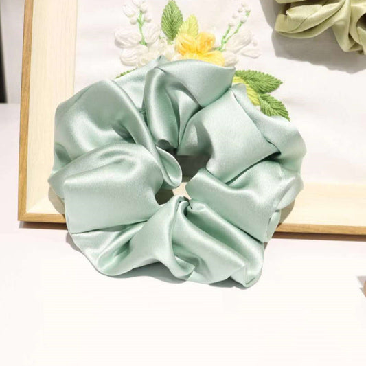 Mint green mulberry silk scrunchie with ruched folds resting against a wooden-framed floral embroidery piece