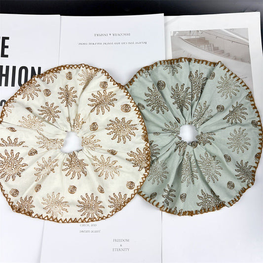 Two fabric scrunchies with sunburst-patterned embroidery—one cream-colored and one mint green—rest on a white surface with printed text and a grayscale photo in the background.