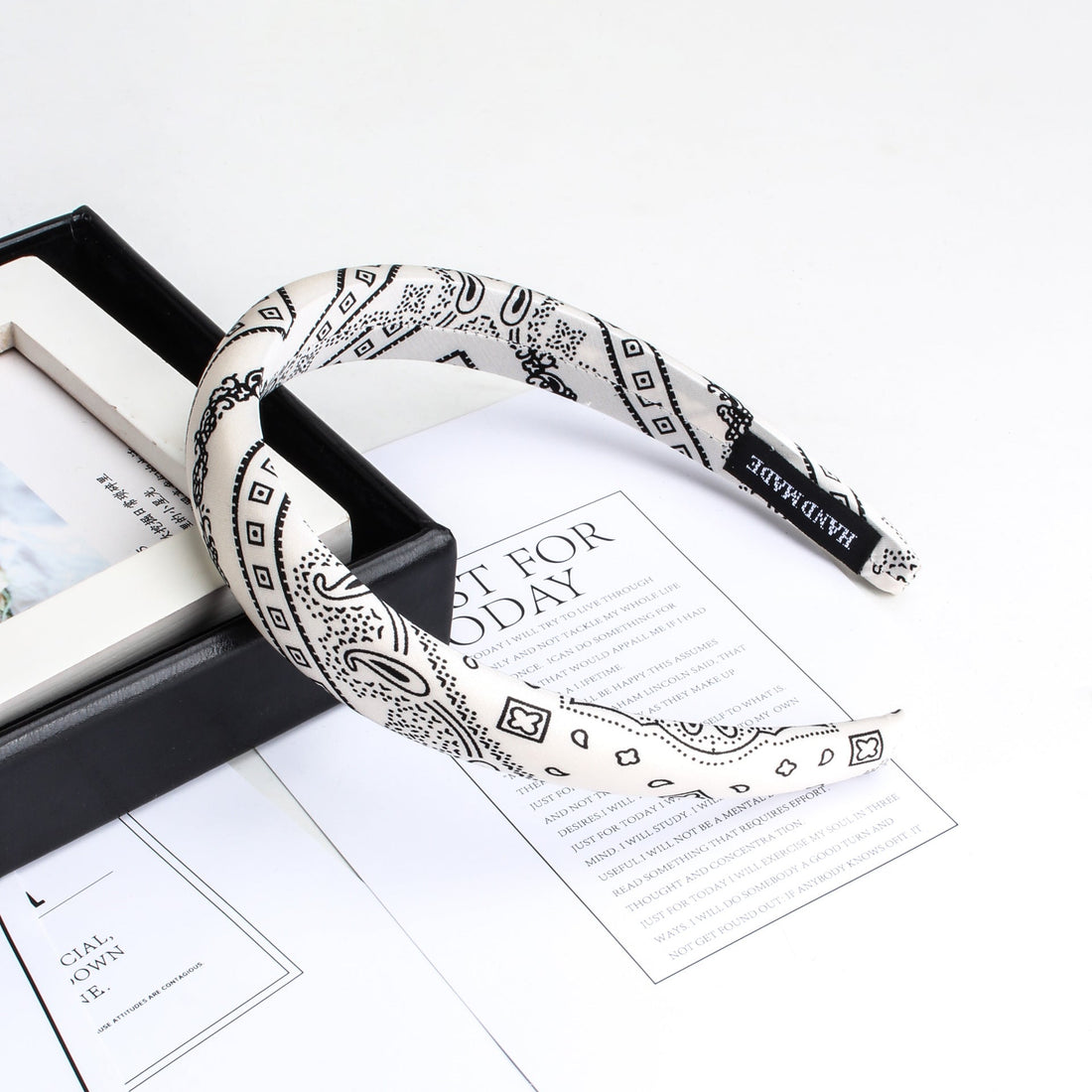 White headband with black paisley and geometric pattern on stack of papers and black box, handmade tag visible