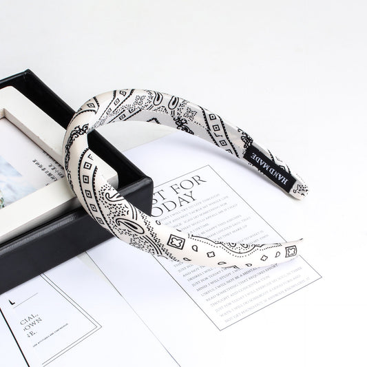 White headband with black paisley and geometric pattern on stack of papers and black box, handmade tag visible