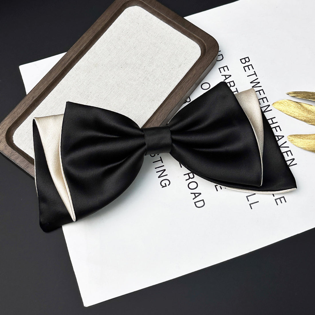 Black satin bow tie with cream-colored inner lining resting on a white paper with printed text next to a wooden-framed rectangular object