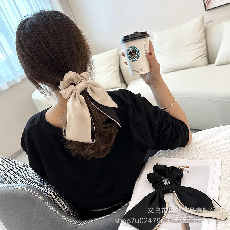 Person with dark hair viewed from behind, wearing a beige and black fabric bow hair tie while holding a coffee cup, with a matching black and beige bow hair tie resting on a magazine on a table in the foreground.