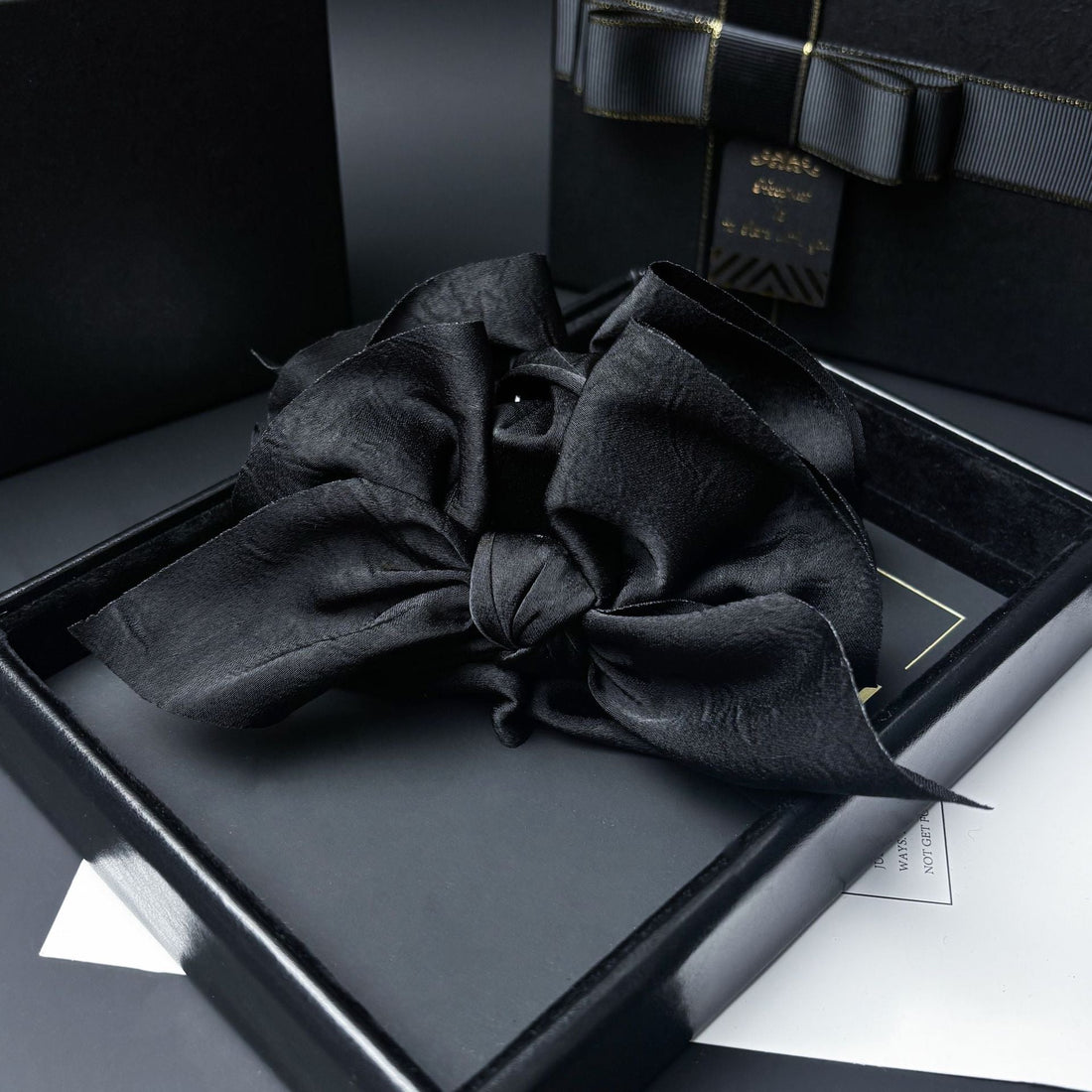 Close-up of a black satin bow with a dark central bead, resting on a dark gray lining inside a black gift box.