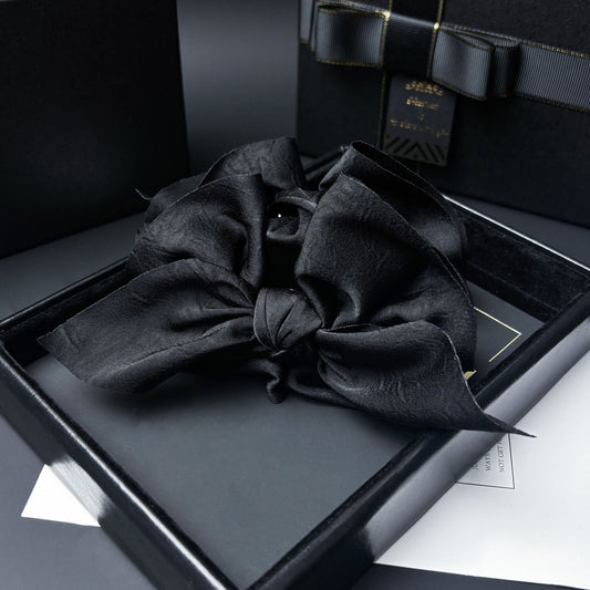 Close-up of a black satin bow with a dark central bead, resting on a dark gray lining inside a black gift box.